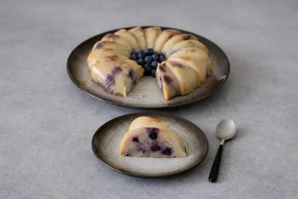 Flan aux myrtilles en forme de couronne, entamé avec une part servie à l’avant sur une petite assiette, révélant une texture dense et fondante, sur une table grise avec une cuillère à côté.