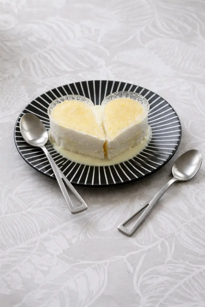 Dessert crémeux en forme de cœur, coupé en deux, présenté sur une assiette noire avec deux cuillères, texture fondante et nappage léger