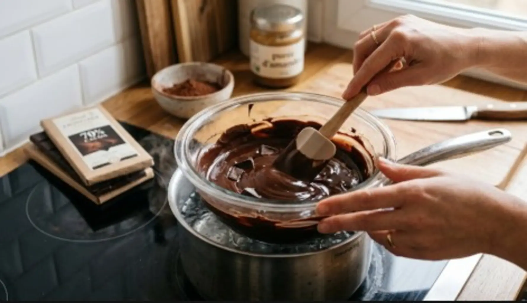 Une main utilise une spatule en silicone pour remuer du chocolat noir en cours de fonte dans un bol en verre placé au-dessus d'une casserole d'eau frémissante pour un bain-marie. La texture du chocolat est onctueuse, lisse et brillante avec quelques morceaux encore entiers. En arrière-plan, sur un plan de travail lumineux, on aperçoit une tablette de chocolat noir 70 %, un pot de purée d'amandes et un petit bol de poudre de cacao.