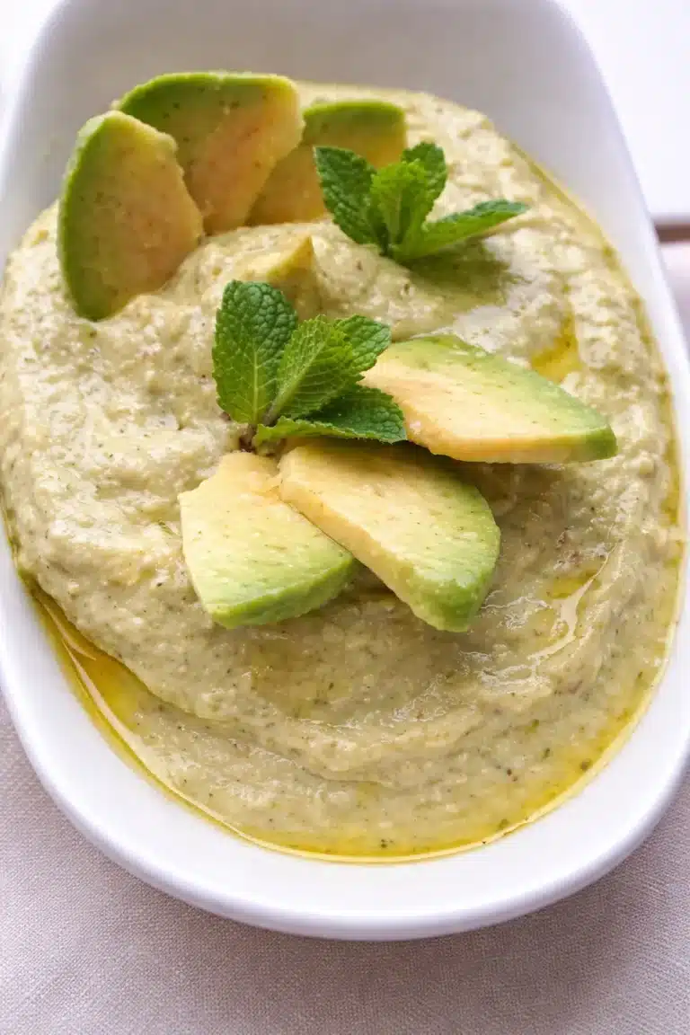 Houmous avocat crémeux en gros plan dans un bol blanc avec tranches d’avocat, menthe fraîche et filet d’huile d’avocat