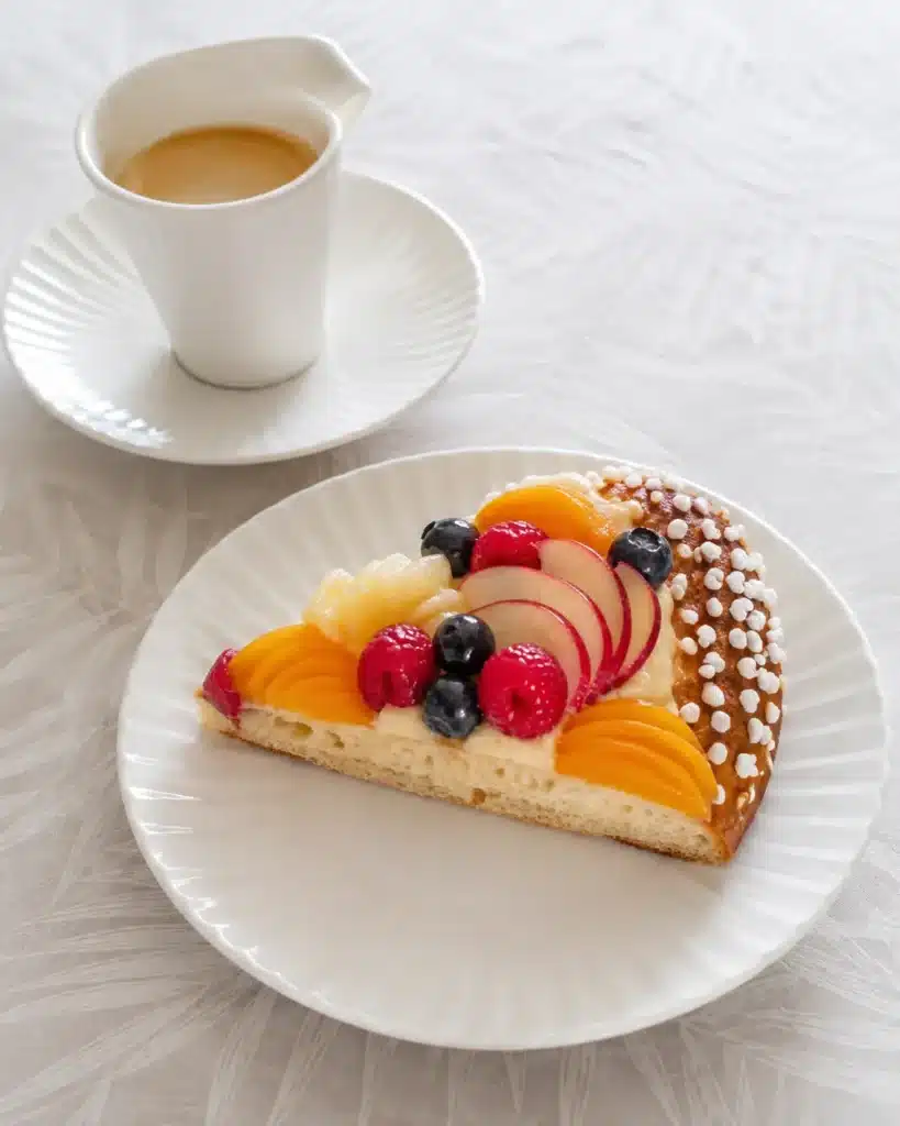 Part de fougasse catalane garnie de crème catalane et de fruits de saison, servie dans une assiette avec une tasse de café.